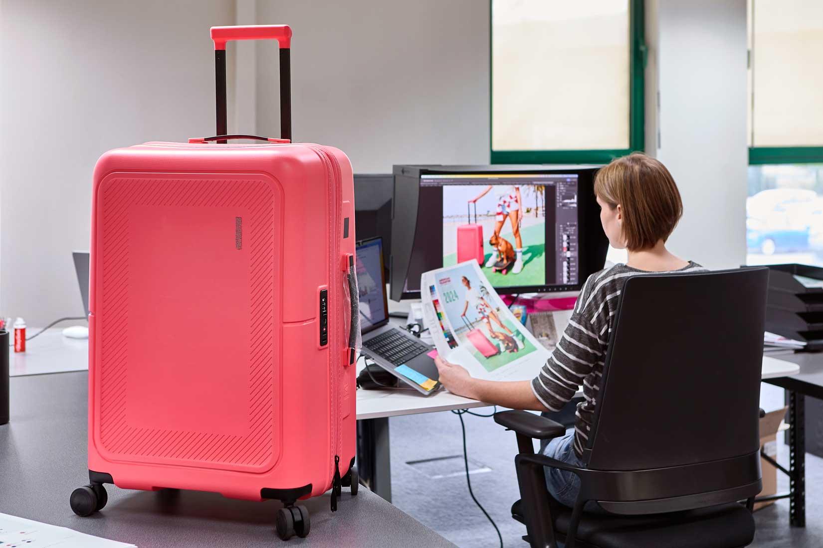 Brandle - Beeldwerking fotografie van American Tourister Roze American Tourister reiskoffer in een kantooromgeving, naast een designer die beeldmateriaal controleert op een computerscherm.