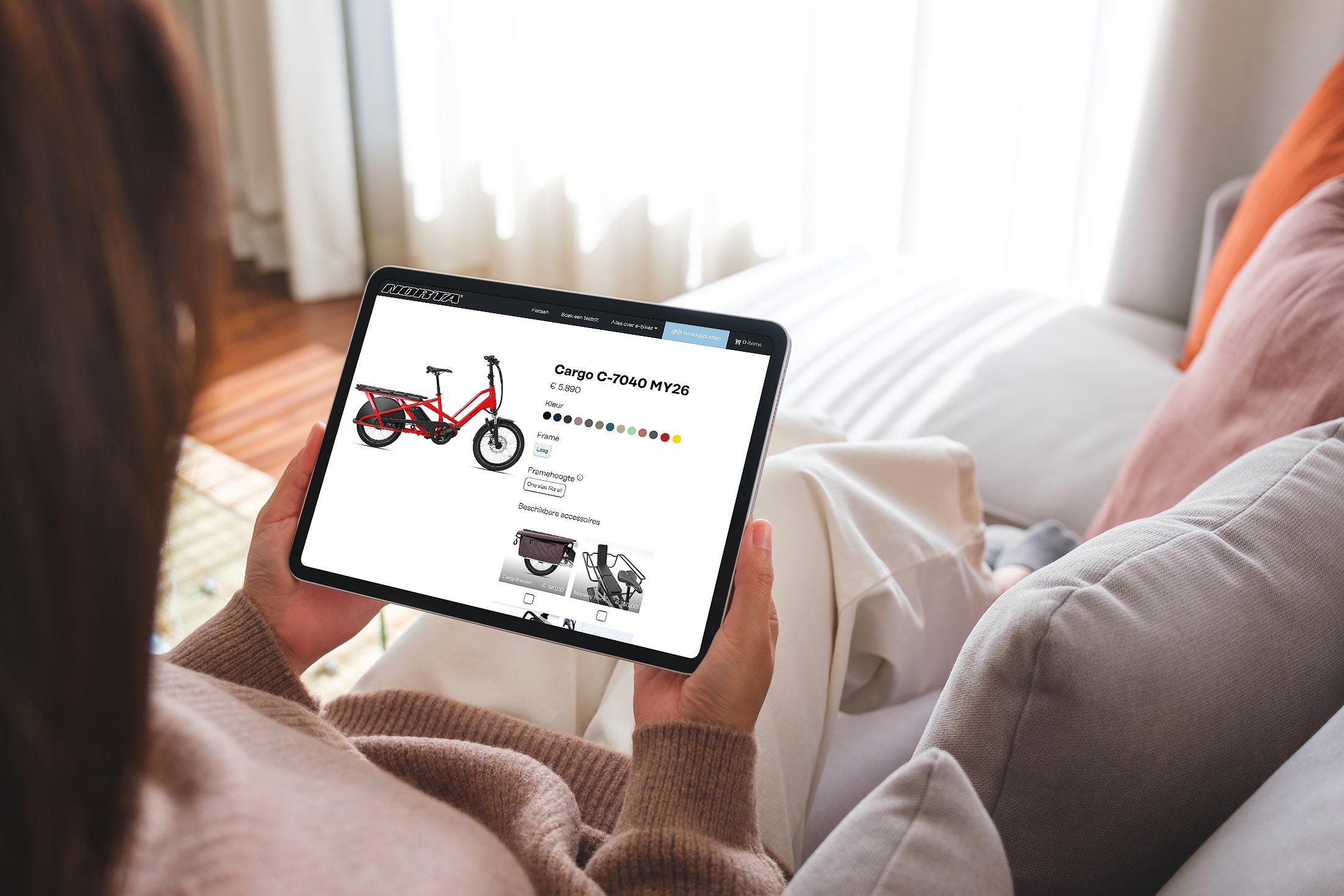 Brandle - Website met configuratie norta.be op tablet Persoon bekijkt op een tablet de productpagina van een rode elektrische cargofiets op de Norta-website, zittend op een bank.