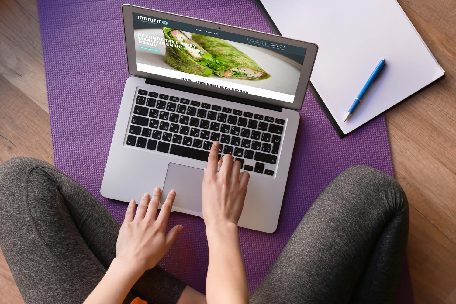 Brandle - Webshop Ecwid van Tastyfit Persoon werkt op laptop met Tastyfit-website, zittend op yogamat met notitieboek en pen ernaast.