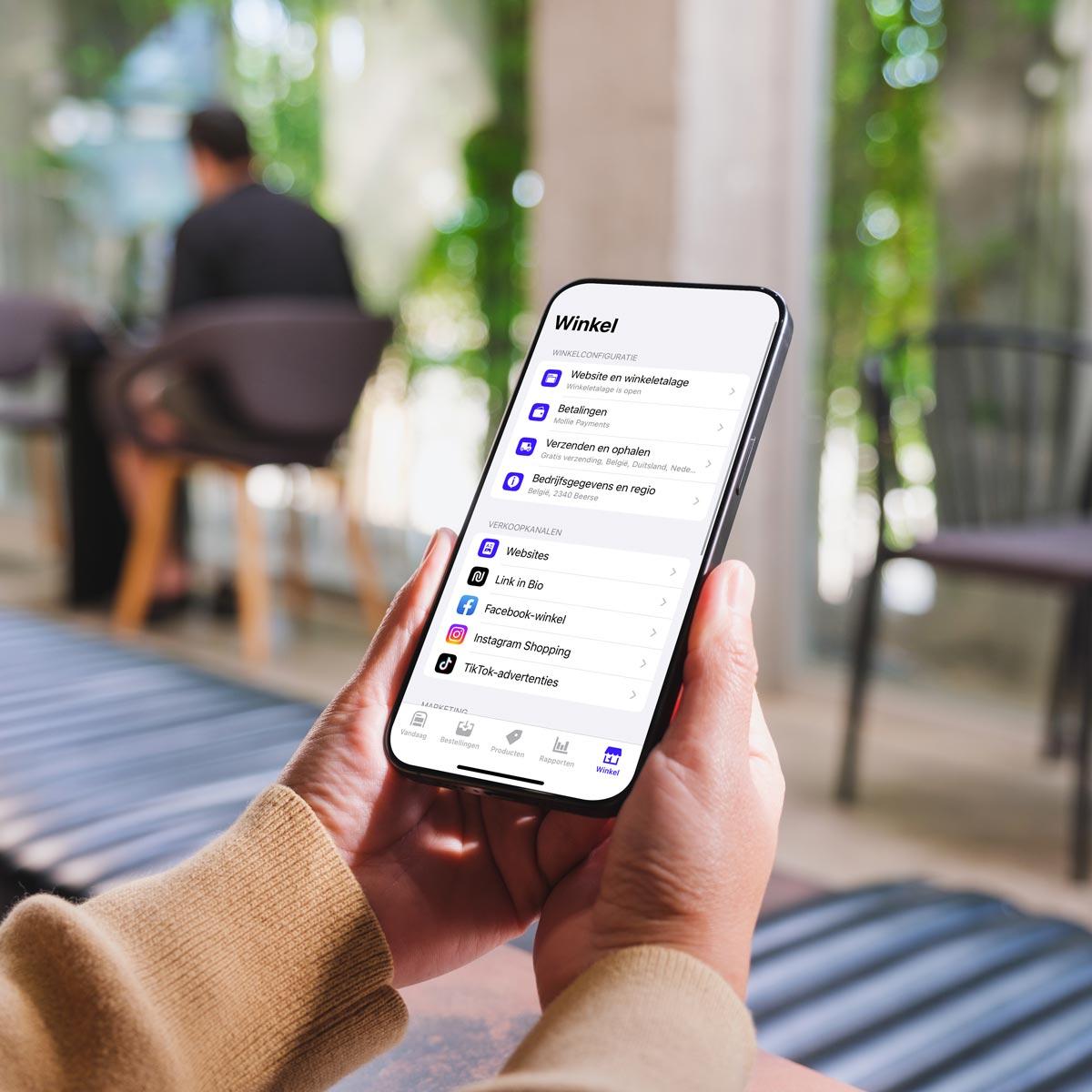 Brandle - Webshops beheren met een app Handen houden een smartphone vast met de Ecwid winkel-app, waarop instellingen voor webshop, betalingen en verkoopkanalen zichtbaar zijn.